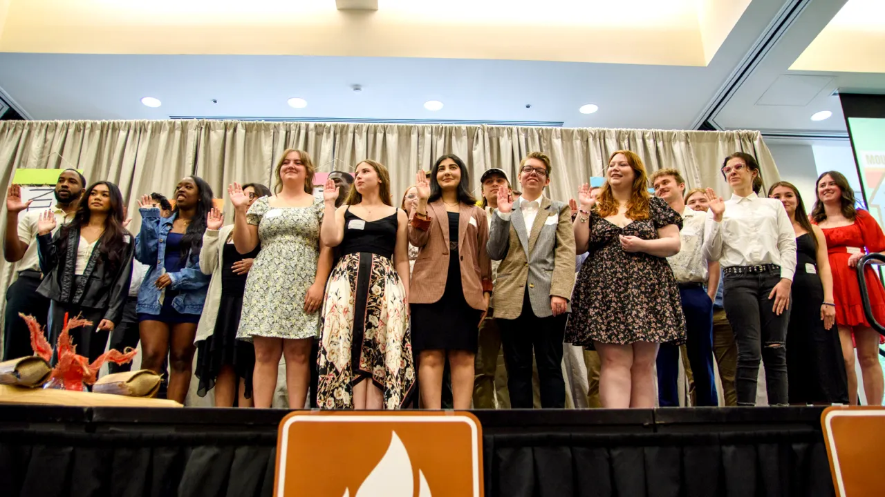 A group of people stand on a stage with their right hands raised, participating in a formal ceremony. They are dressed in a mix of business and casual attire, with a backdrop of light-colored curtains.