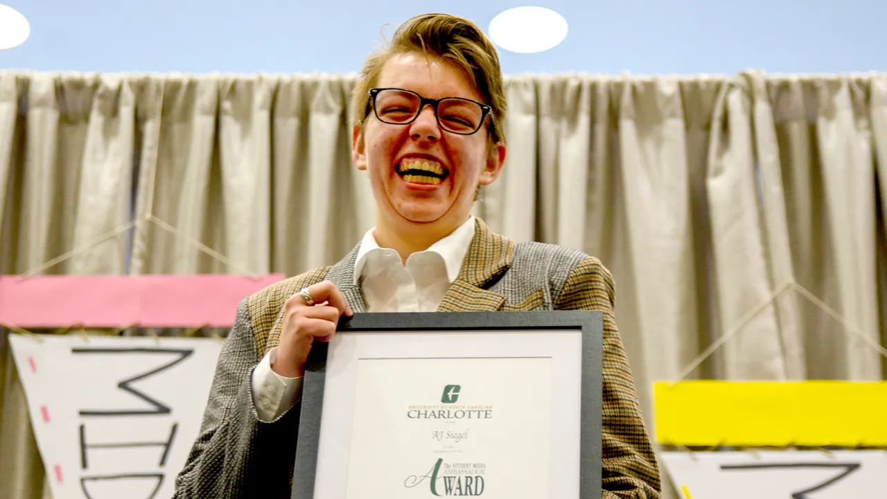 A person wearing glasses and a suit is smiling broadly while holding a framed certificate. A curtain and banners are visible in the background.