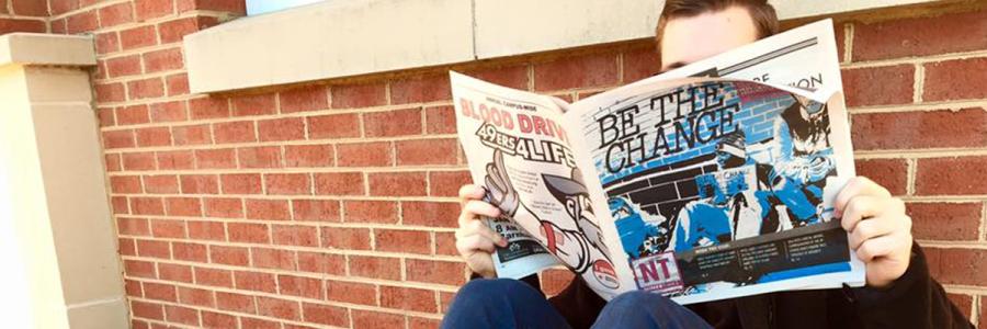 A male student reading a issue of Niner Times, sitting against a brick wall