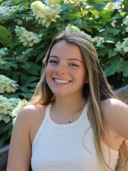 A smiling person with long hair outdoors, with white flowers in the background.