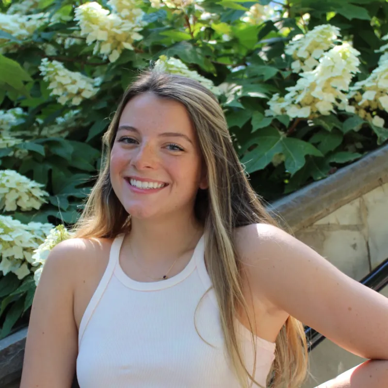 A smiling person with long hair outdoors, with white flowers in the background.