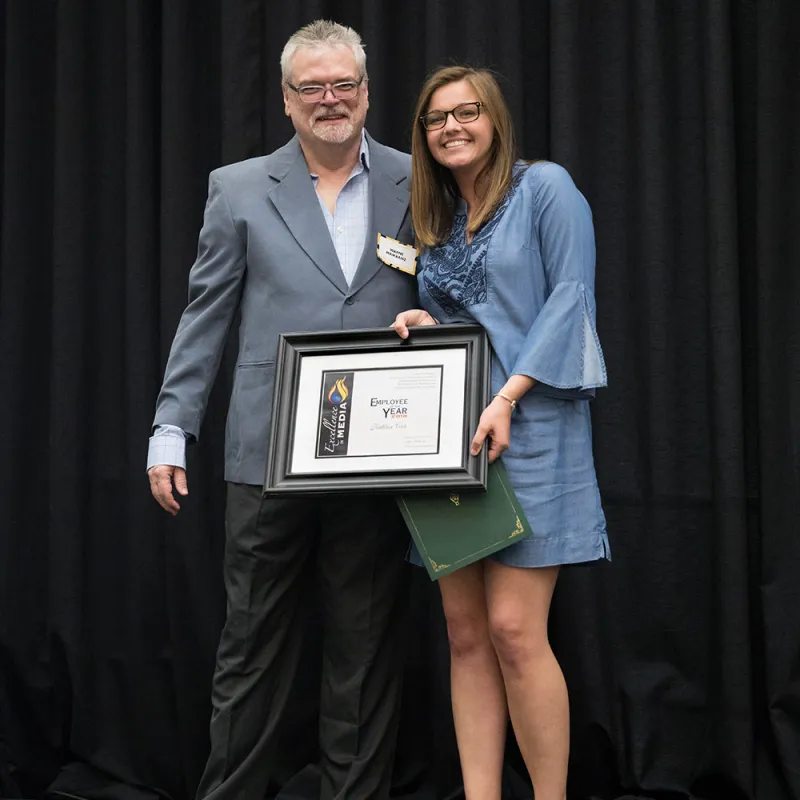 Media Advisor, Wayne Maikranz, presenting student, Kathleen Cook, with Employee of the Month Award. Against a dark backdrop.